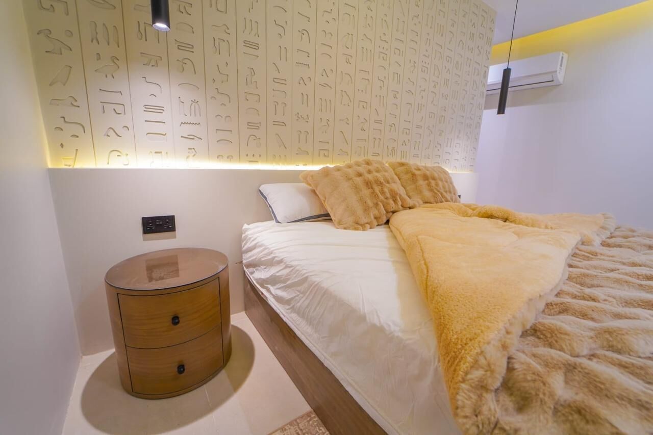 A bedroom with a bed, a bedside table, a wall with hieroglyphs, and an air conditioning unit.