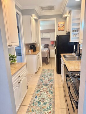 A compact kitchen with white cabinetry, a black refrigerator, a countertop with a coffee maker, and a patterned rug on the floor. - Cozy 2-bedroom Bungalow near Disney, Universal, SeaWorld, Legoland (Kissimmee)