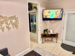 A living room with a TV, a wooden decorative wall piece, a small wooden table with a plant, and a door. - Cozy 2-bedroom Bungalow near Disney, Universal, SeaWorld, Legoland (Kissimmee)