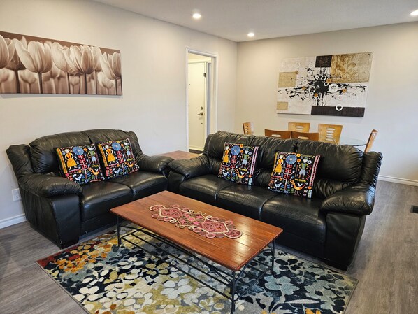 Una sala de estar con un sofá de cuero negro, cojines decorativos de colores, una mesa de centro de madera y una alfombra estampada.