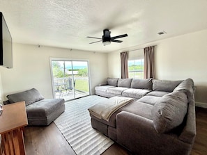 A modern living room with a grey sectional sofa, a small wooden table, a grey armchair, a ceiling fan, and a sliding glass door leading to an outdoor area.