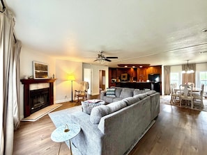 A living room with a sofa, a small round table, a fireplace, a ceiling fan, and a kitchen area with wooden cabinets and a TV.