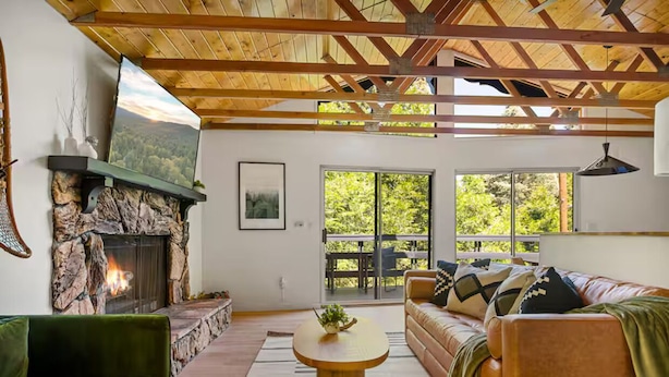 Una sala de estar con chimenea de piedra, un sofá verde, una mesa de centro de madera y un ventanal con vistas a zonas verdes.
