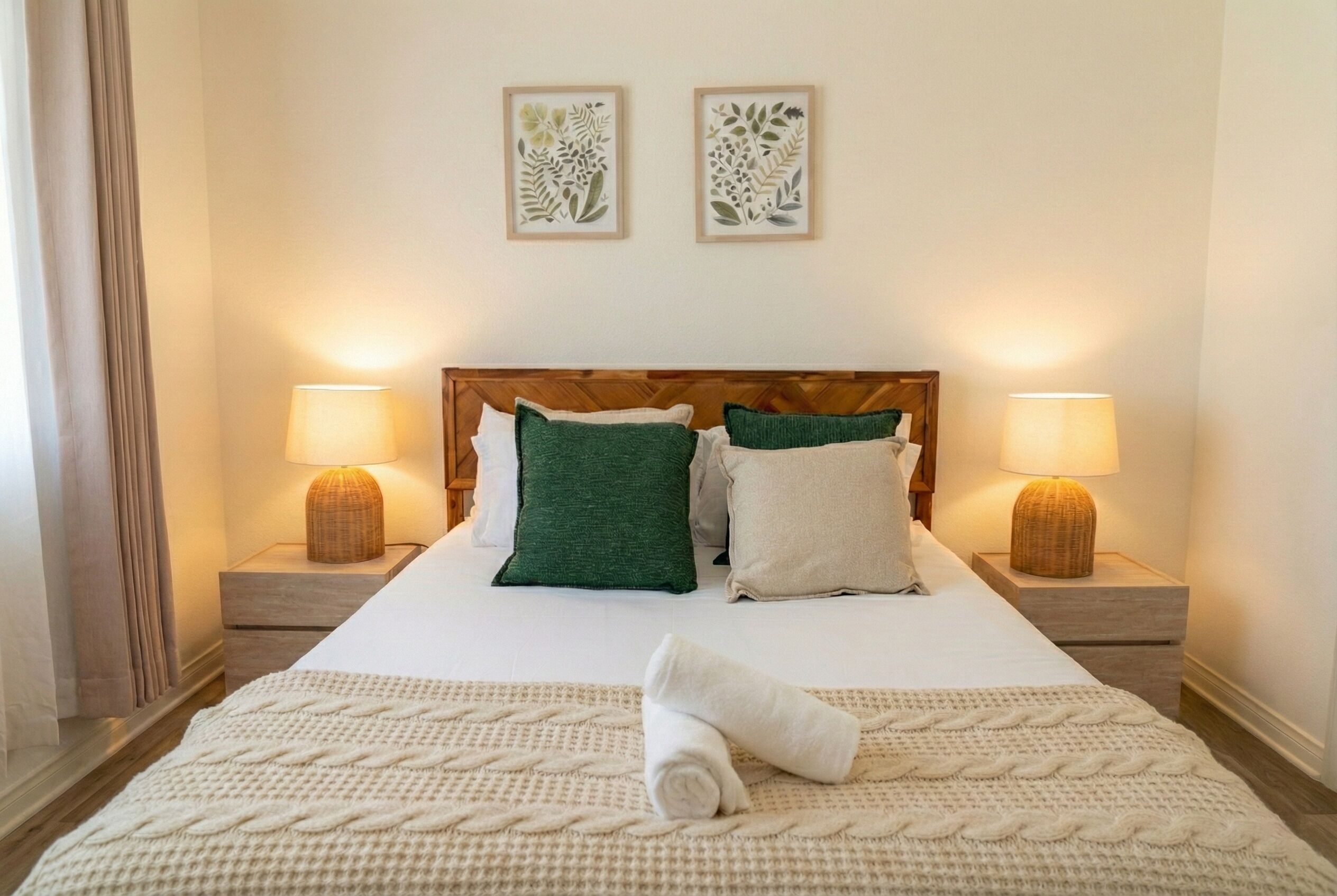 Un lit bien fait, recouvert d’une couverture blanche et agrémenté d’une serviette roulée, flanqué de deux lampes de chevet, dans une chambre dotée d’une tête de lit en bois et de deux reproductions botaniques encadrées au mur.