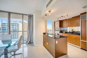 A modern kitchen with wooden cabinets, a central island, and a dining area with a glass table and chairs. - Icon Brickell - 36th Floor - Ocean & City Views (Miami)