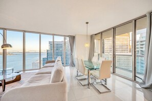 A modern living room with a glass dining table, white sofas, and large windows offering a city view. - Icon Brickell - 36th Floor - Ocean & City Views (Miami)