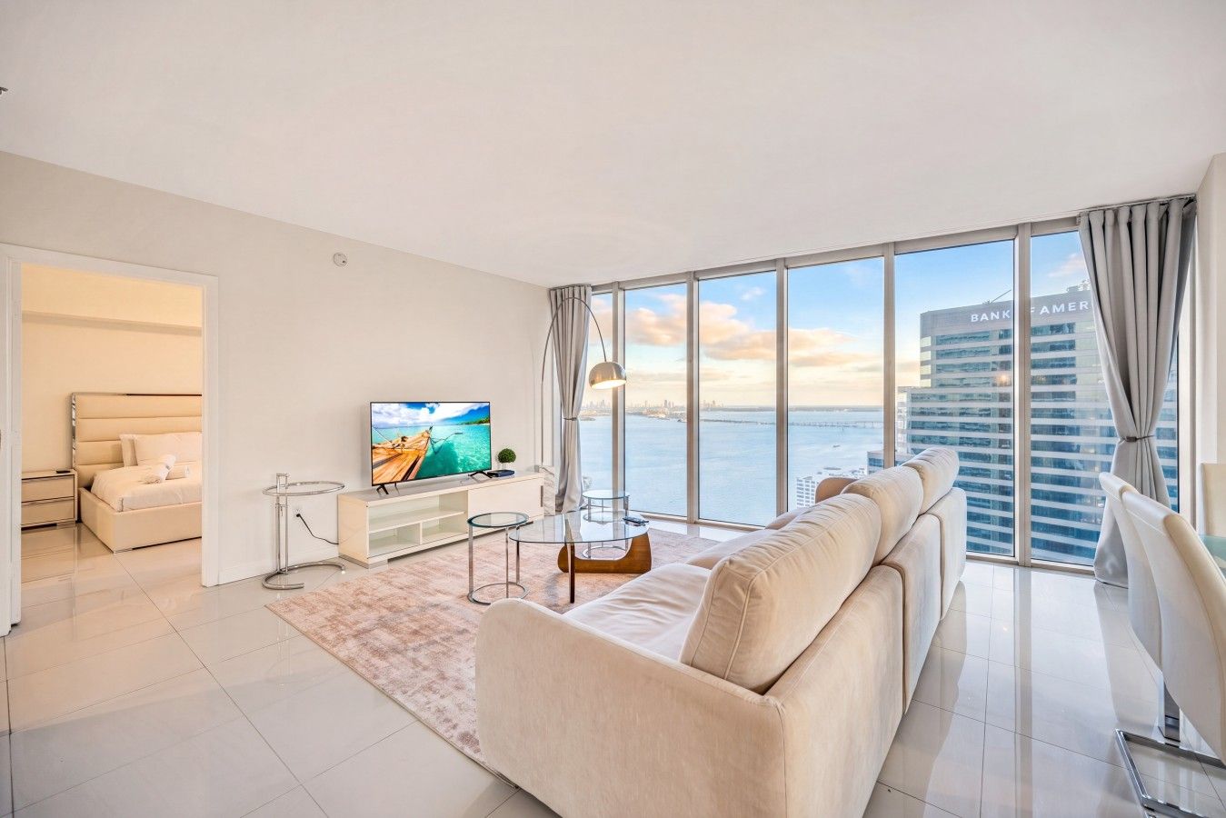 A modern living room with a large TV, a sofa, a glass coffee table, and a view of the water and buildings.