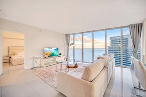 A modern living room with a large TV, a sofa, a glass coffee table, and a view of the water and buildings. - Icon Brickell - 36th Floor - Ocean & City Views (Miami)