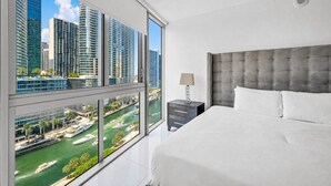 Habitació d'hotel moderna amb un llit gran, una tauleta de nit i vista a edificis alts i un port esportiu.