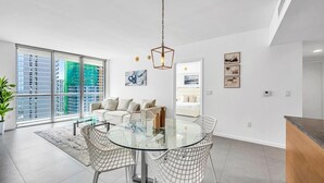 Una sala de estar moderna con una mesa de comedor de vidrio, sofás blancos y vistas a edificios altos.