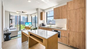 A modern kitchen with wooden cabinets, a dining area with a table, and a view of the cityscape through large windows.