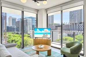 Ein modernes Wohnzimmer mit großem Fernseher, einer Couch, einem Couchtisch und einem Sessel, mit Blick auf die Stadt durch große Fenster.