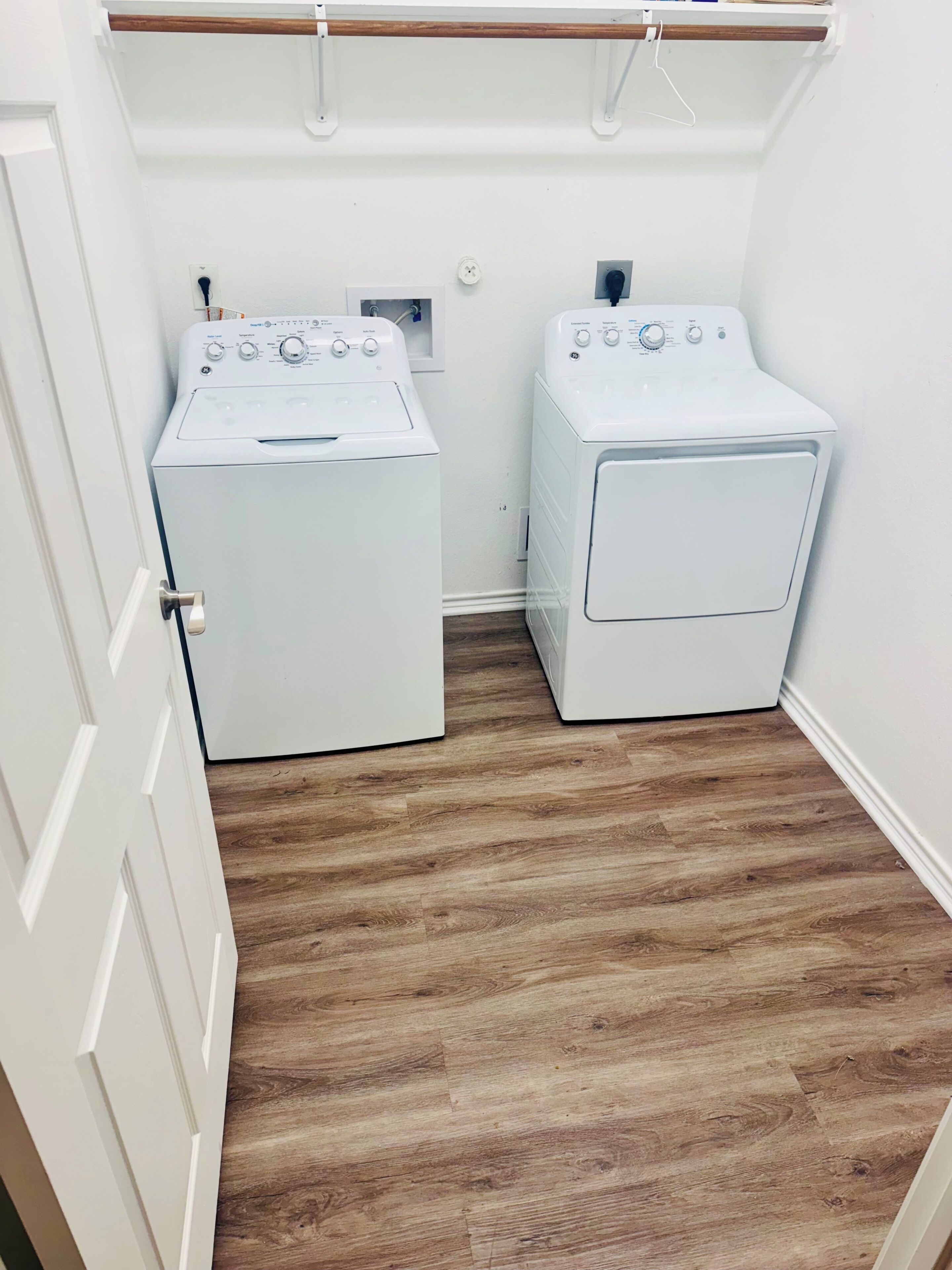 Une buanderie équipée de deux machines à laver et de deux sèche-linge à chargement frontal, tous deux de couleur blanche, avec un plancher en bois et une porte blanche.