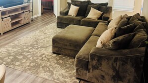 A living room with a sectional sofa, a TV on a wooden stand, a ceiling fan, and a patterned rug.