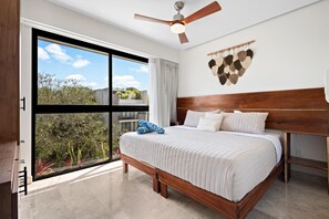 Ein Schlafzimmer mit großem Fenster, Deckenventilator, Holz-Kopfteil und Bett mit weißer Bettwäsche.