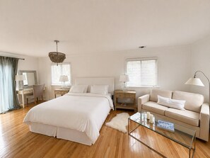 A bedroom with a large bed, a glass coffee table, a sofa, a desk, and a chandelier. - Peconic Bay Cottage (Hampton Bays)