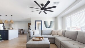 Una sala moderna con un sofá modular, un ventilador de techo y una cocina al fondo.