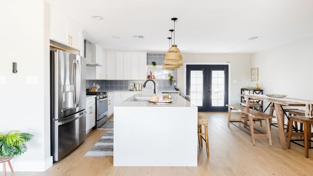 Una cocina moderna con armarios blancos, electrodomésticos de acero inoxidable, una isla central y un comedor con mesas y sillas de madera.
