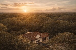 A house with a tiled roof surrounded by dense vegetation, a clear sky, and the sun setting in the background. - Luxury 5BR Villa - Hacienda Pinilla - Pool, Beach Club, Breakfast, House Keeping (Tamarindo)