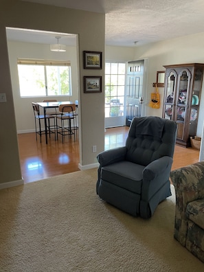 Una sala de estar con un sillón gris, un sillón estampado, una zona de comedor con mesa y sillas, y un aparador con puertas de cristal.