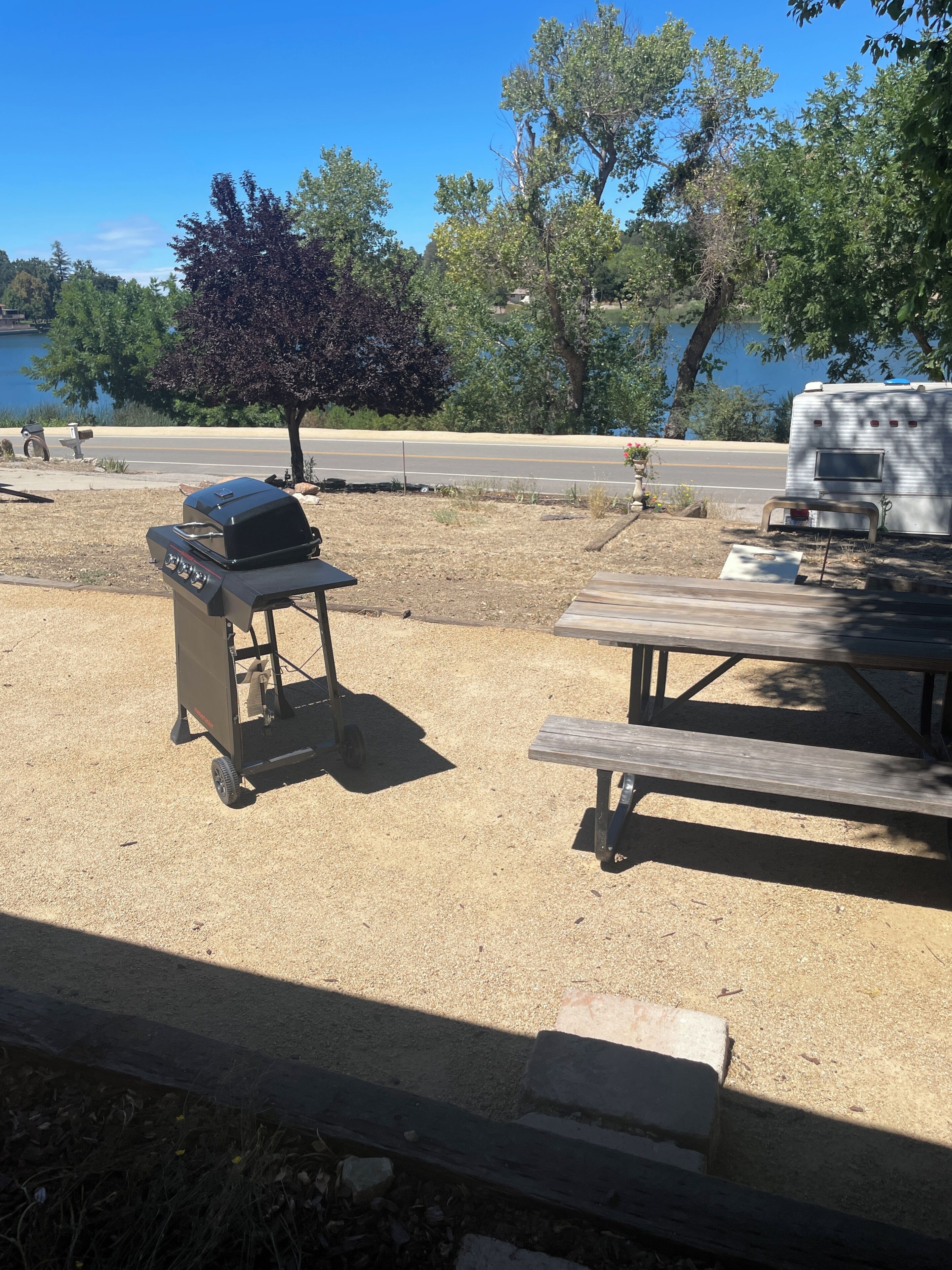 A portable grill and a picnic table in a grassy area with trees and a road in the background.