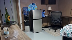 Ein Hotelzimmer mit Schreibtisch aus Holz, einem Kühlschrank, einem Wasserspender, einem Fernseher, einem Sessel und einem Bett mit schwarzem Teppich.