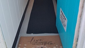 Ein Flur mit einer schwarzen Matte, die zu einer Tür mit dem Schild 'Welcome' führt, einer Lampe und einem Gestell mit Gegenständen.