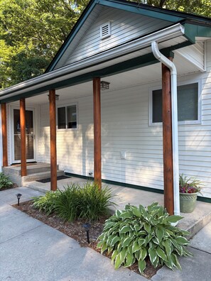 A white house with a porch supported by wooden posts, a front door, and a small window. - The Cottage at Midpoint. Charming 3 bedroom retreat near Higgins Lake. (Roscommon)