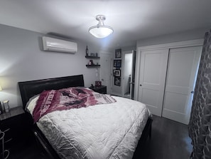 A bedroom with a bed, an air conditioner, a ceiling light, a door, and a wall-mounted shelf.