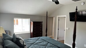 Un dormitorio con cama, cómoda, ventilador de techo, televisor y ventana con persianas.