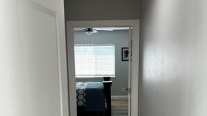 Un pasillo que conduce a un dormitorio con cama, ventilador de techo y ventana con persianas.