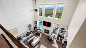 A spacious living room with a fireplace, a TV, and a view of a mountainous landscape through large windows. - Ultimate Family Retreat: VIEWS, Sport Court, Theater, Pool, Sauna, Game Zone! (Heber City)