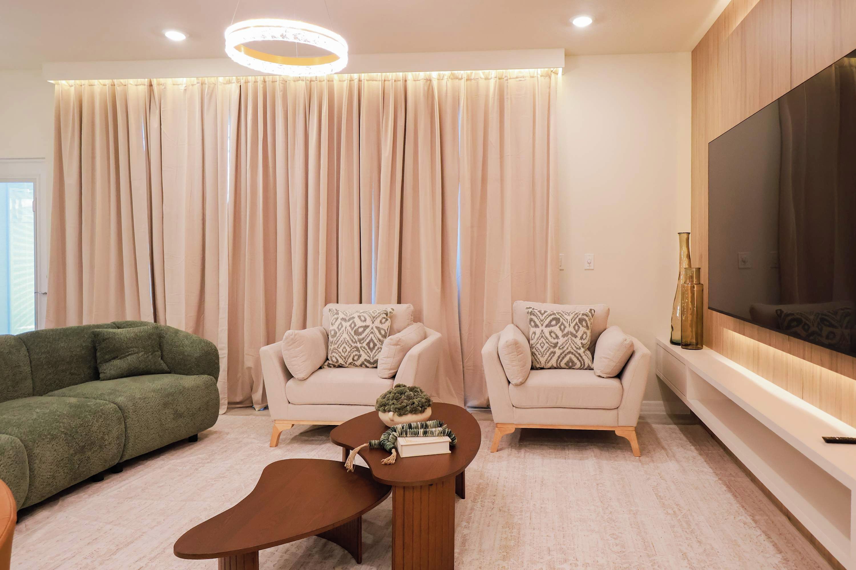 A modern living room with a green sofa, two beige armchairs, a round coffee table, and a large flat-screen TV mounted on the wall.