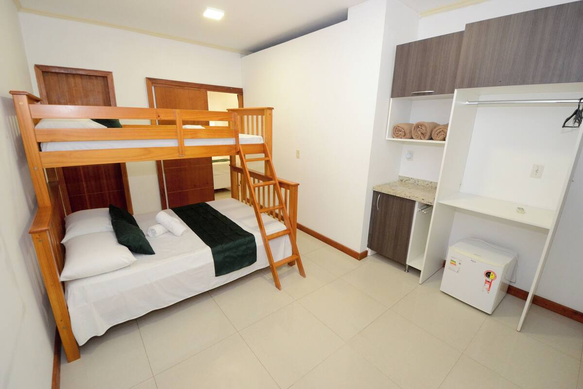 A bunk bed with a ladder, a small desk, a wardrobe, and a mini fridge in a room with white walls and tiled flooring.