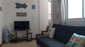A living room with a TV, a sofa, a surfboard, and wall decorations including a 'Beach' sign and a 'I need vitamin sea' sign. - Cozy 2-bedroom apartment in charming Tamarindo with AC (Tamarindo)