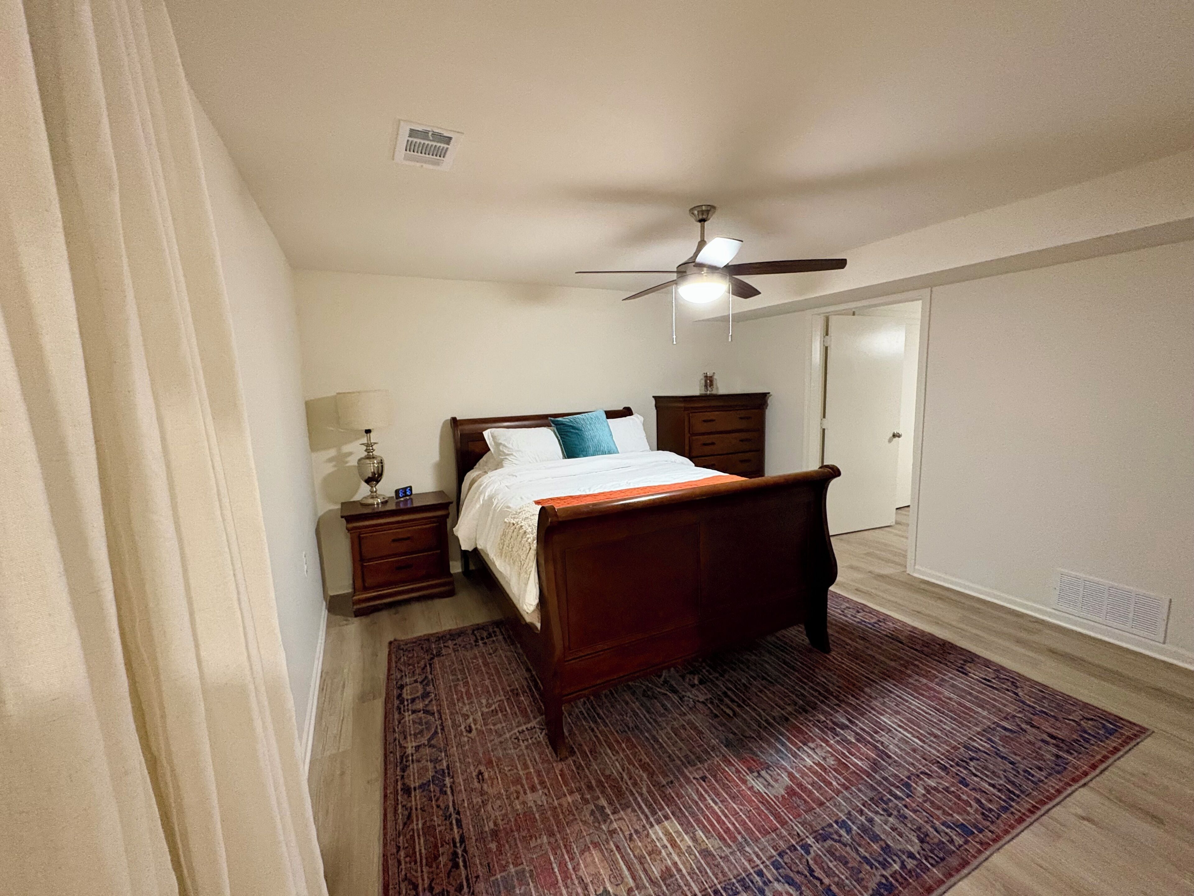 Une chambre à coucher comprenant un lit, une table de chevet, une commode, un ventilateur de plafond et une porte.