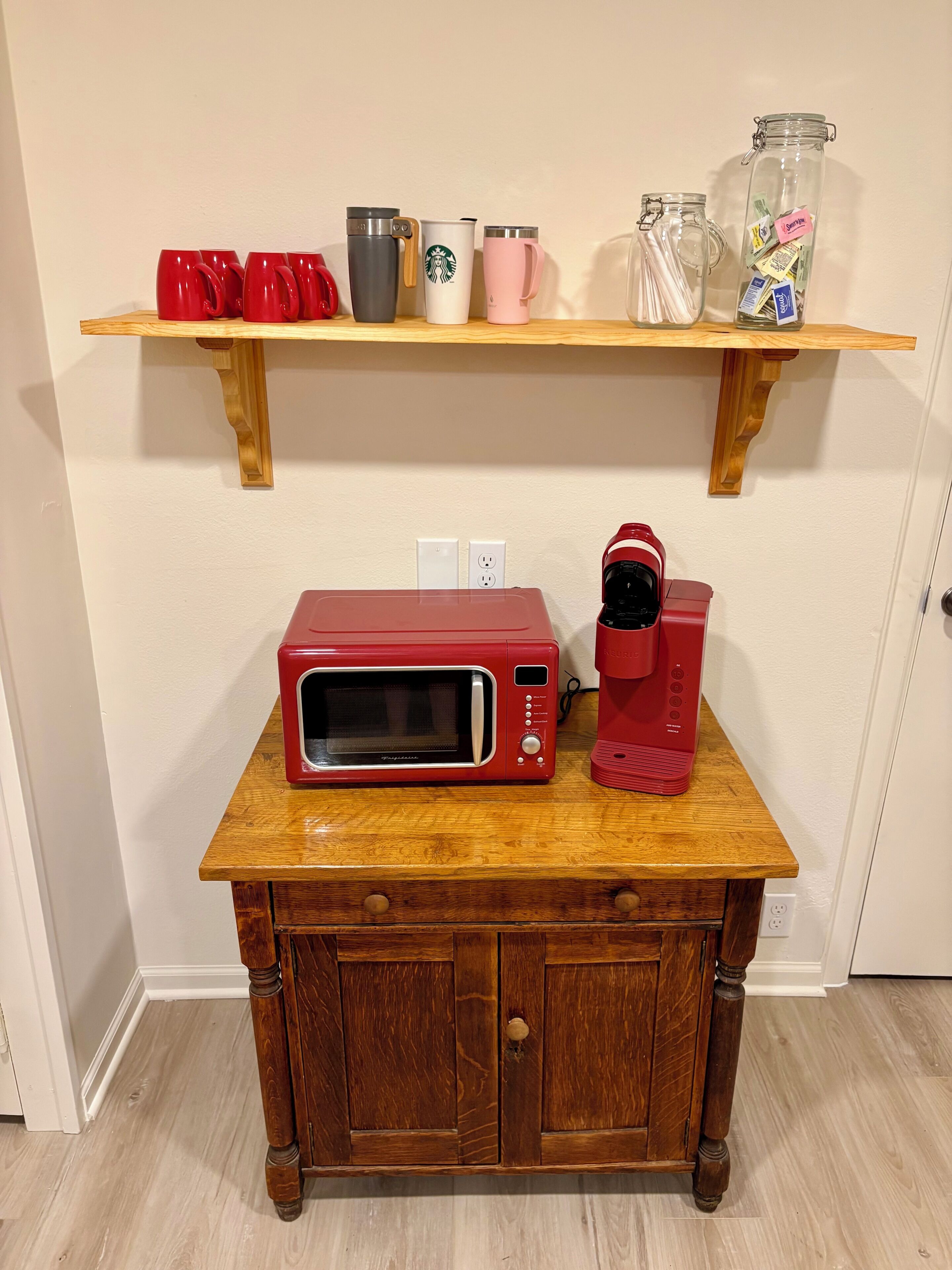 A kitchen counter with a red microwave, a red coffee maker, and a shelf above holding various mugs and jars.