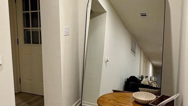 Een gang met houten vloer, een ronde tafel met een spiegel, en een deur naar een andere kamer.