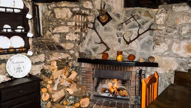 Una chimenea de piedra con fuego, una cabeza de ciervo montada encima y una repisa con platos y tazas.
