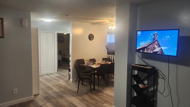 A living room with a dining area, a TV mounted on the wall, and a hallway leading to another room.