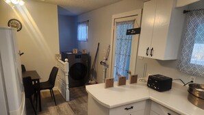 A compact kitchen with white cabinets, a built-in washing machine, a small dining area with a table and chairs, and a window with patterned curtains.