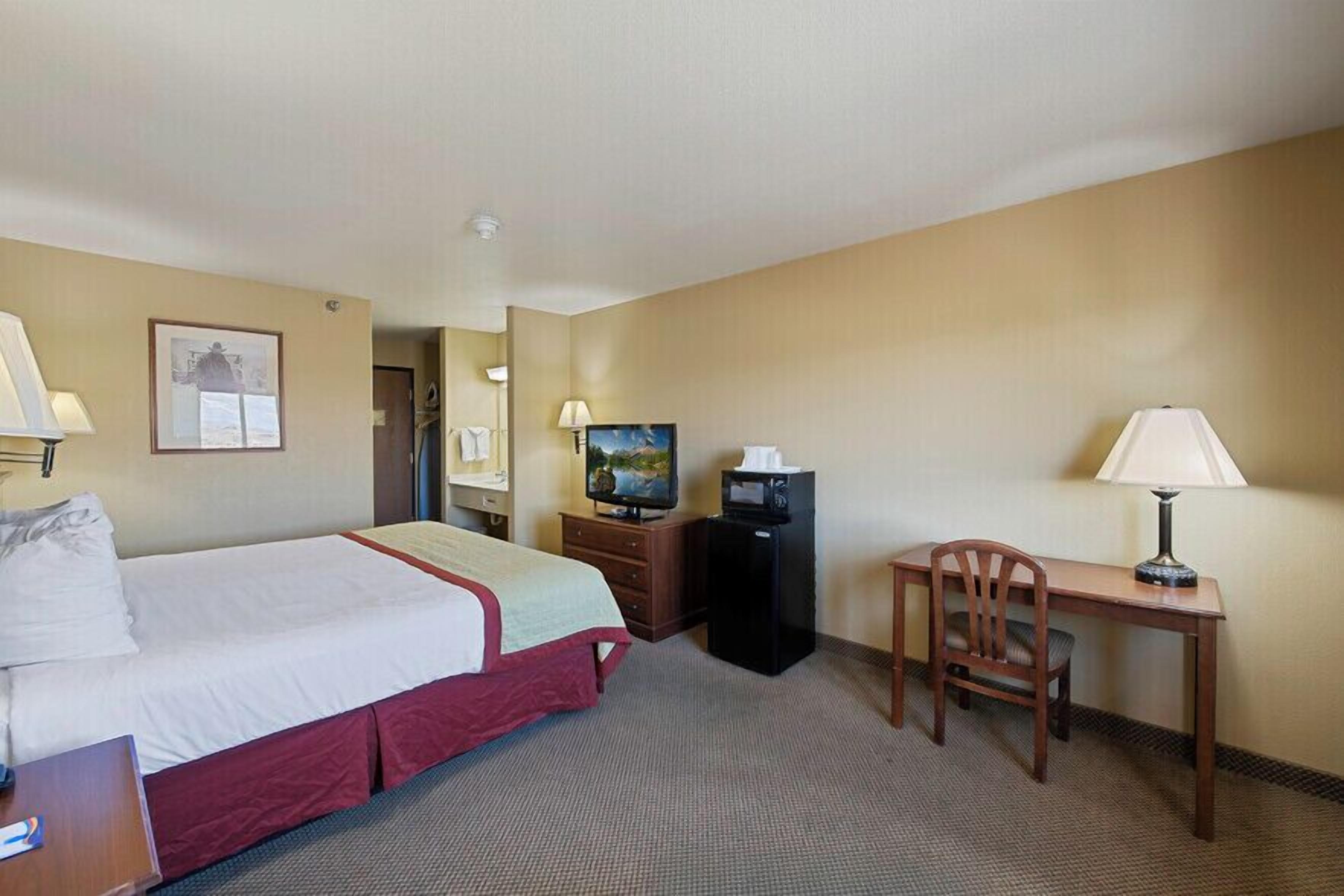 A hotel room with a bed, a desk with a chair, a television, and a small table with a lamp.