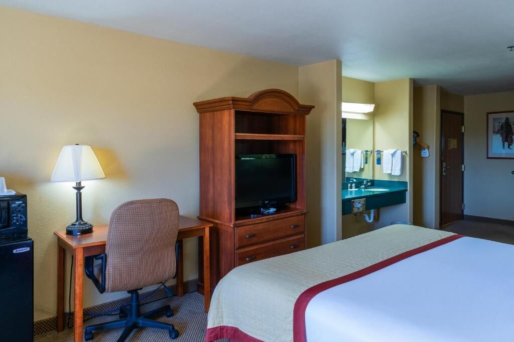A hotel room with a bed, a desk with a lamp, a television, a chair, a dresser, a mirror, and a bathroom with a sink and towel rack.