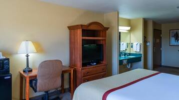 A hotel room with a bed, a desk with a lamp, a television, a chair, a dresser, a mirror, and a bathroom with a sink and towel rack.