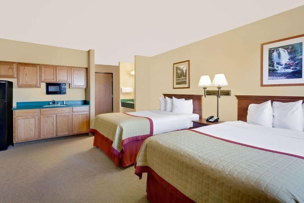A hotel room with two beds, a kitchenette, a bathroom, and a painting on the wall.