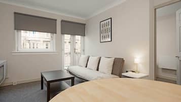 A bedroom with a bed, a bedside table, a lamp, a window with blinds, and a wall with a 'GOOD VIBES ONLY' sign.