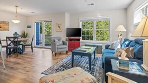 A living room with a sofa, a coffee table, a TV, a dining table with chairs, and a window with a view of greenery. - Bayfront TC Condo · Walk to Beach & Downtown · Free Parking · Sleeps 6 (Traverse City)