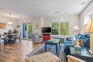 A living room with a sofa, a coffee table, a TV, a dining table with chairs, and a window with a view of greenery. - Bayfront TC Condo · Walk to Beach & Downtown · Free Parking · Sleeps 6 (Traverse City)
