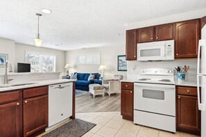 A compact kitchen with white appliances, wooden cabinets, a blue sofa, and a dining area. - Bayfront TC Condo · Walk to Beach & Downtown · Free Parking · Sleeps 6 (Traverse City)
