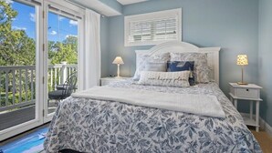 A bedroom with a bed, two bedside tables, a window with blinds, a ceiling fan, and a balcony with a chair. - Bayfront TC Condo · Walk to Beach & Downtown · Free Parking · Sleeps 6 (Traverse City)